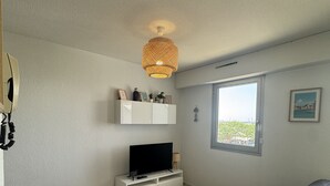 Ett modernt rum med en tv på ett vitt skåp, ett glasbord, en svart soffa, en taklampa av rotting och ett fönster med utsikt.