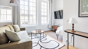 Een moderne woonkamer met een bank, een glazen salontafel, een aan de muur bevestigde televisie en een lamp op een bijzettafeltje.
