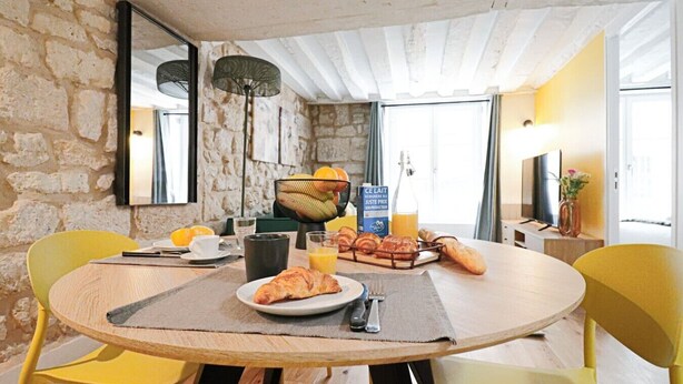Ein Frühstückstisch mit runden Stühlen, einem Steinwandhintergrund, einem Obstkorb, Croissants und einer Orangensaftflasche.