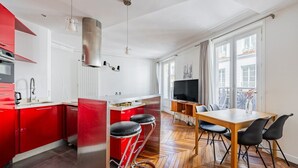 A modern kitchen with red cabinets, a dining area with a wooden table and black chairs, and a TV in the corner. - Flexible Lease – Paris 2 – Montorgueil – 2-bedroom apartment (Paris)