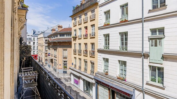 A narrow street with multi-story buildings, balconies, and a storefront with a sign. - Flexible Lease – Paris 2 – Montorgueil – 2-bedroom apartment (Paris)