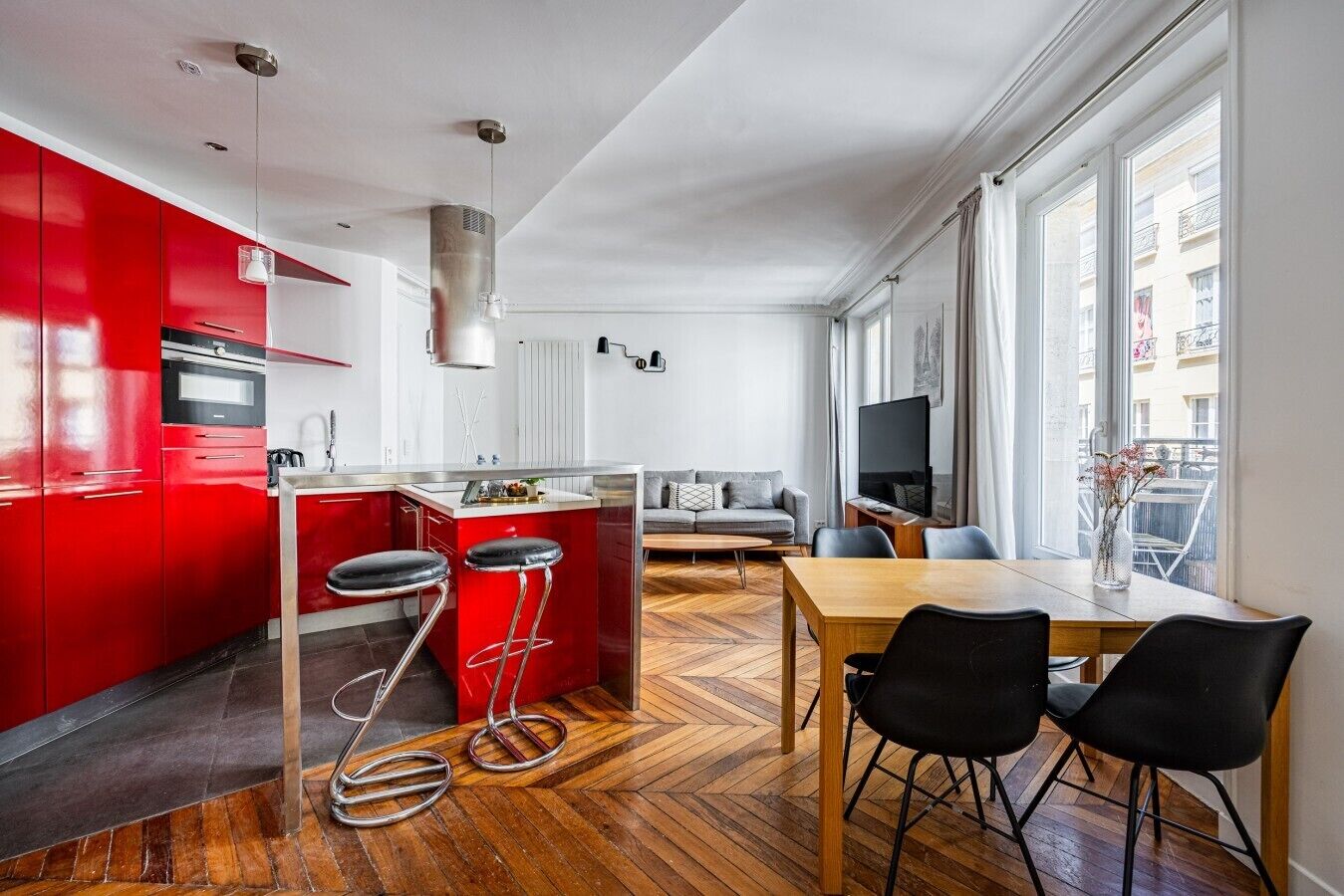 Une cuisine moderne avec des meubles rouges, un îlot central doté de tabourets, un coin repas avec une table et des chaises en bois, et un salon avec un canapé et un téléviseur.