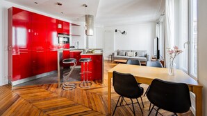 A modern kitchen with red cabinets, a dining area with a wooden table and black chairs, and a living room with a grey sofa. - Flexible Lease – Paris 2 – Montorgueil – 2-bedroom apartment (Paris)
