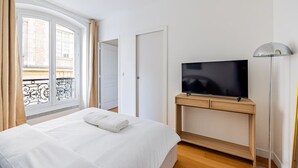 Une chambre d’hôtel avec un lit, un téléviseur posé sur un meuble en bois, une lampe sur pied et une fenêtre avec des rideaux.