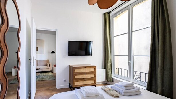 Quarto de hotel com cama, cómoda de madeira, televisão e uma janela grande com cortinas.