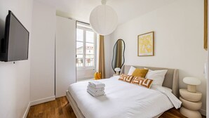 A hotel room with a bed, a TV, a window with curtains, a wall-mounted mirror, and a bedside lamp. - 3. Apartment - 2 people - 1 bedroom - Mouffetard - Sorbonne (Paris)