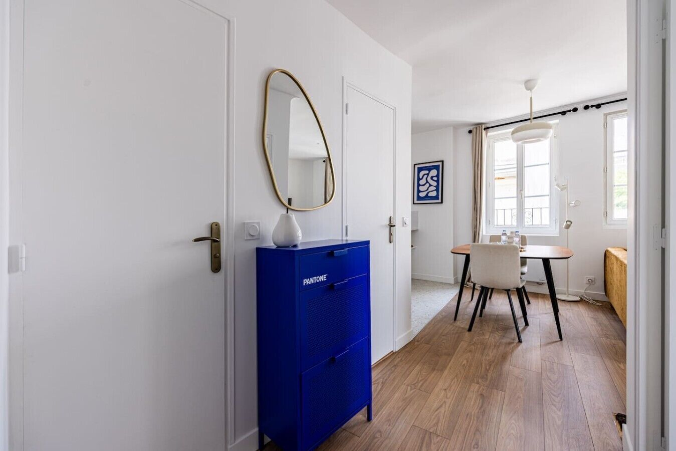 Een hal met een blauwe kast, een spiegel, een eethoek met een tafel en stoelen, en een raam met gordijnen.