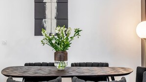 Een ronde eettafel met een vaas witte bloemen, omringd door zwarte stoelen met kussentjes, voor een witte muur met een zwart-wit abstract schilderij.