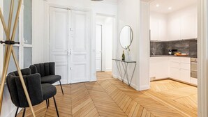 Een modern interieur met een visgraatparketvloer, een zwarte fauteuil, een glazen console tafel met een spiegel, en een keuken met witte kasten en een donkere achterwand.