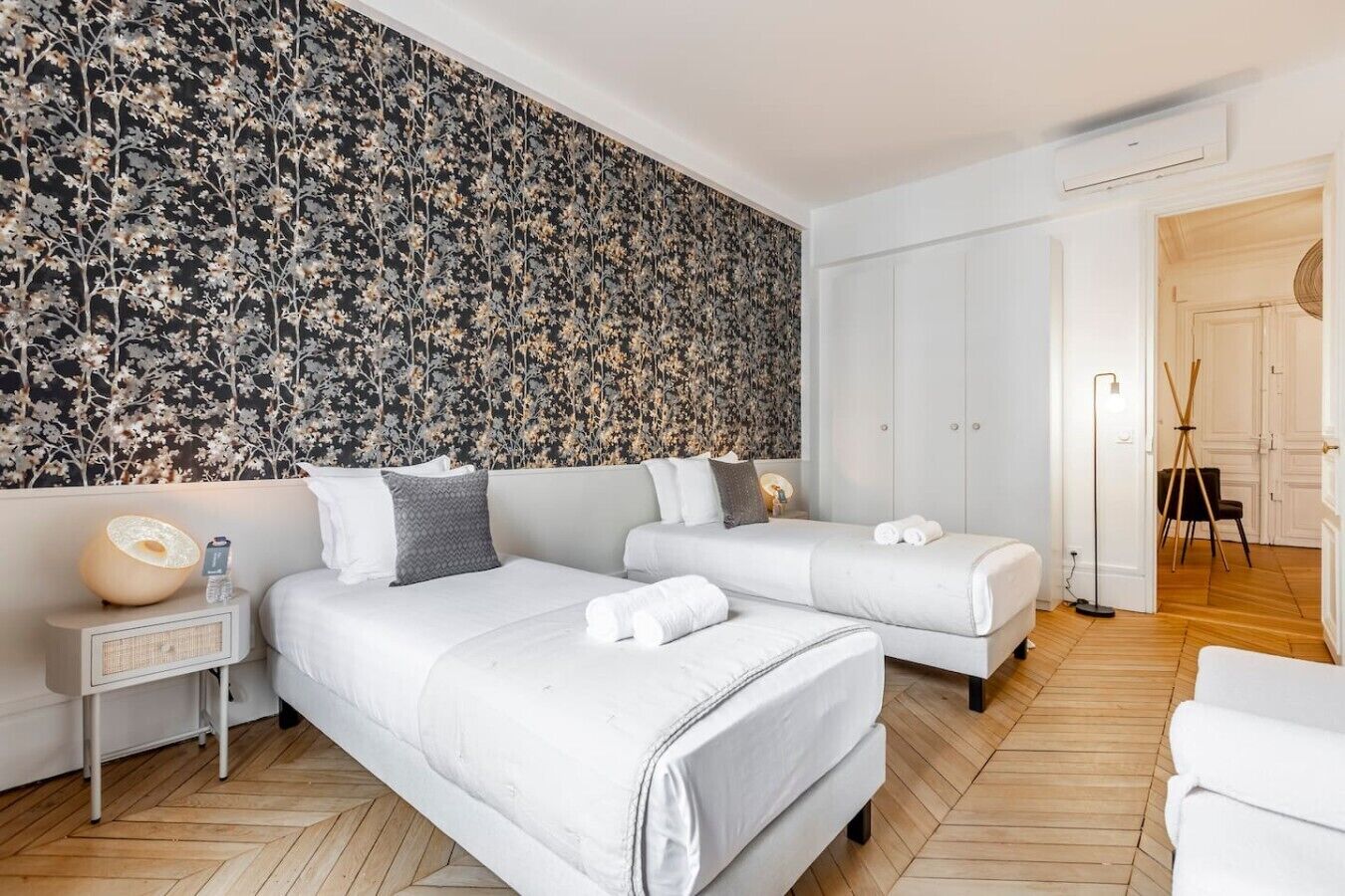 Una habitación de hotel moderna con dos camas, una pared con estampado floral grande, una mesita de noche con una lámpara y suelo de madera.