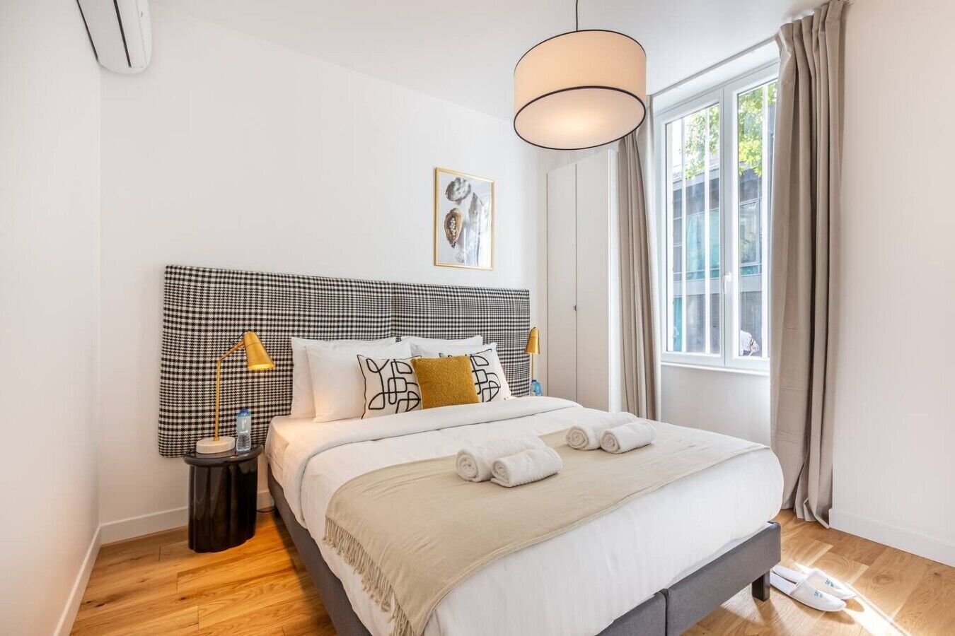 Een netjes ingerichte hotelkamer met een geruit hoofdeinde, een groot raam met gordijnen, een bedlamp en een lijst met een afbeelding aan de muur.