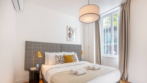 Quarto de hotel bem arrumado com cabeceira xadrez, uma janela grande com cortinas, abajur e um quadro na parede.