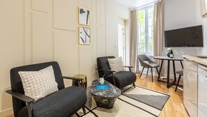 Uma sala de estar moderna com duas poltronas pretas, uma mesa redonda pequena, uma televisão de tela plana e um espaço de jantar com mesa e cadeiras.
