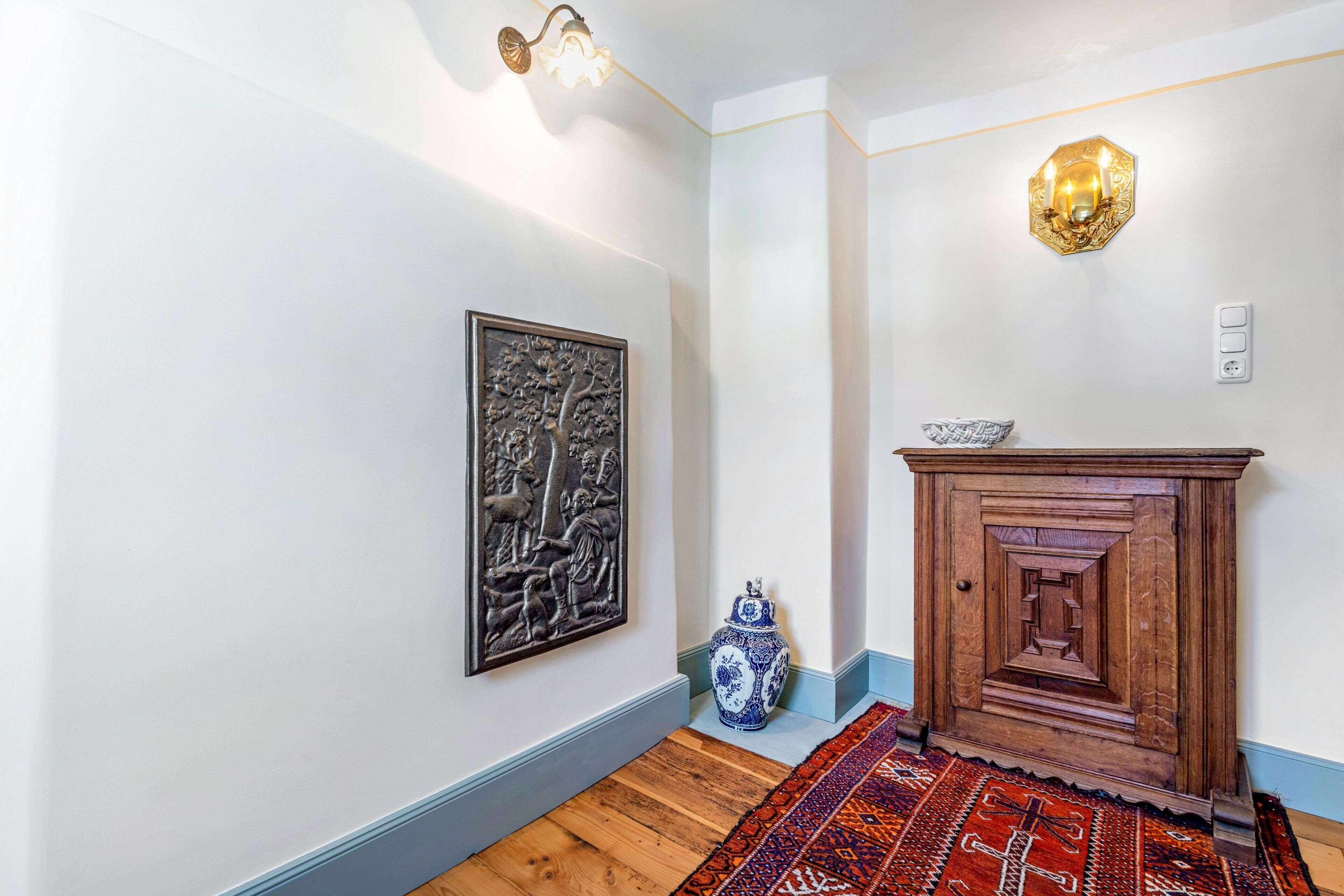 Un couloir avec un luminaire fixé au mur, un élément décoratif mural, un meuble en bois et un tapis à motifs.