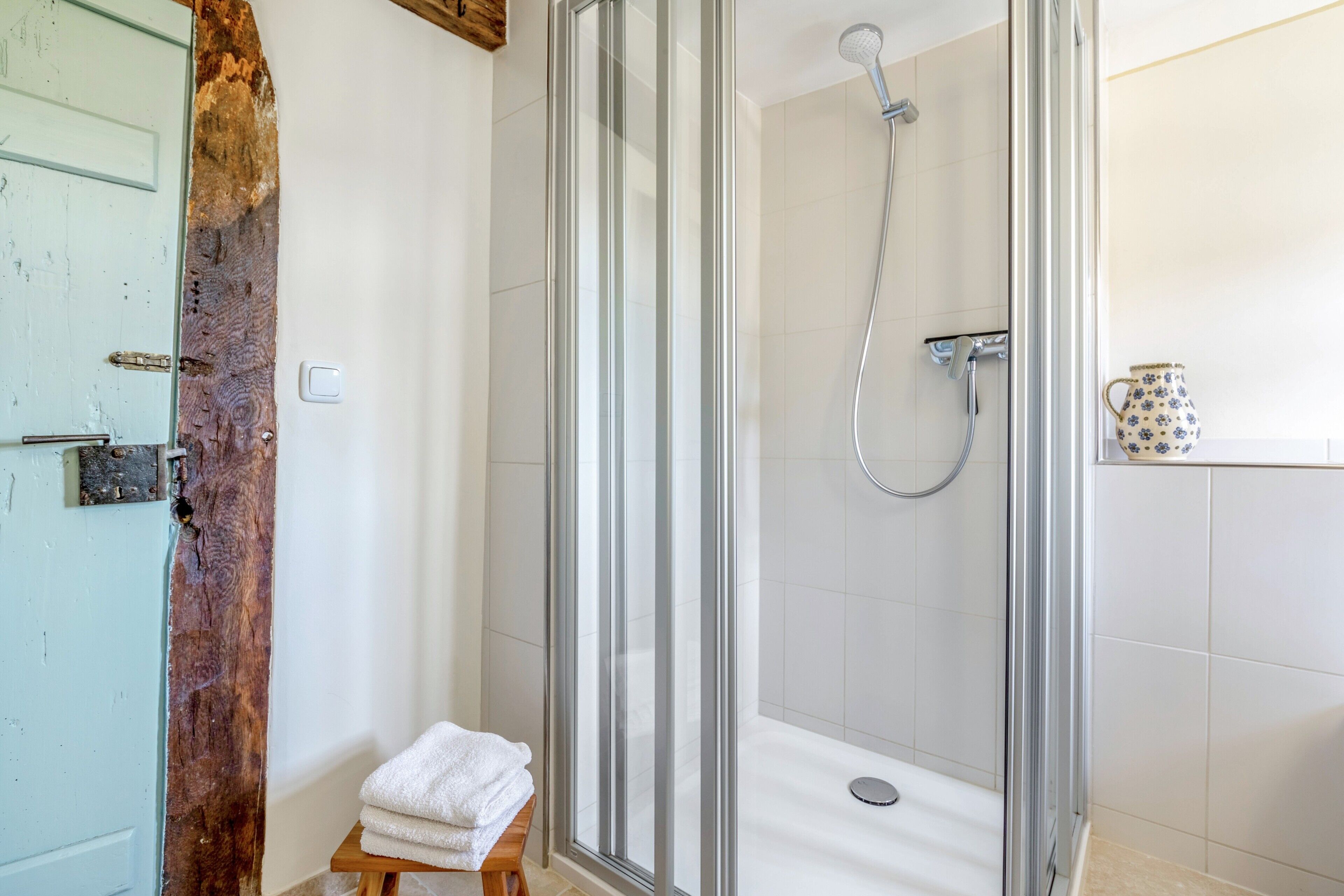 A glass-enclosed shower with a handheld showerhead, a white towel on a wooden stool, and a tiled wall.