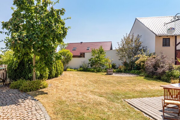 Propiedad residencial con camino de entrada pavimentado, terraza de madera y jardín con diversas plantas y un pequeño árbol.