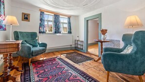 A room with two green upholstered chairs, a wooden floor, a patterned rug, a window with curtains, and a painting on the wall.
