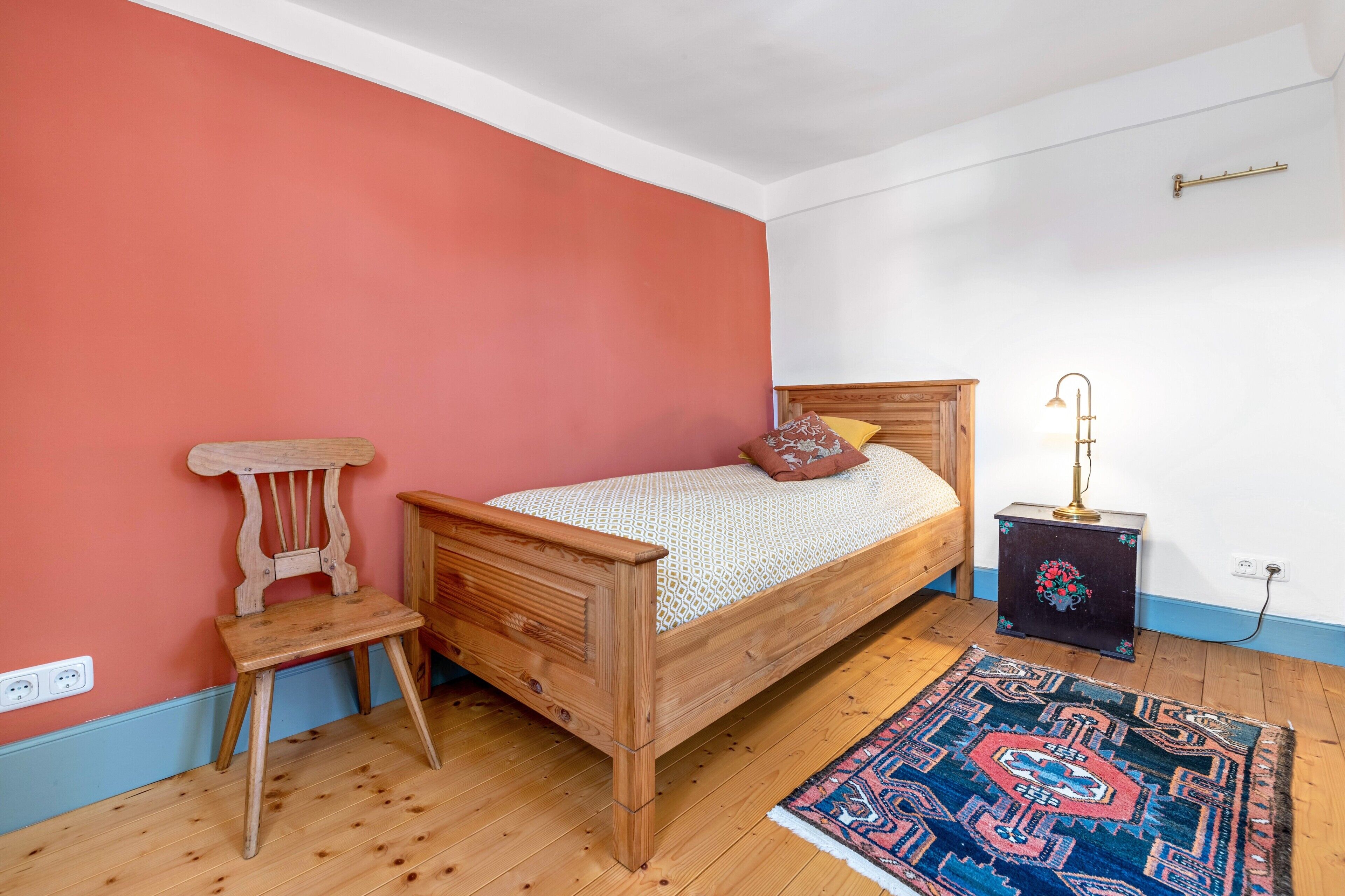 Ein Schlafzimmer mit einem hölzernen Bett, einem Nachttisch, einem Sessel, einem Teppich und einer Steckdose.