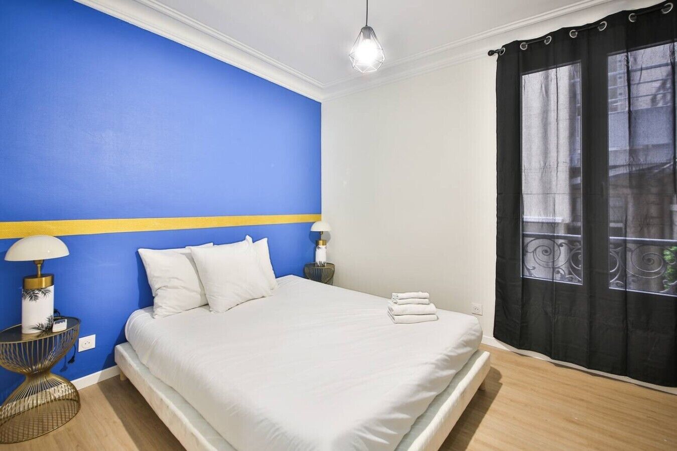 Une chambre avec un mur d’accent bleu, un lit avec du linge de lit blanc, deux lampes de chevet et une fenêtre avec un rideau noir.