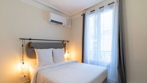 Une chambre à coucher comprenant un lit, un climatiseur fixé au mur, une fenêtre avec des rideaux et un luminaire.