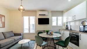 Un espace de vie compact comprenant un canapé, une table de salle à manger ronde avec des chaises, un coin cuisine équipé d’appareils électroménagers et un téléviseur fixé au mur.