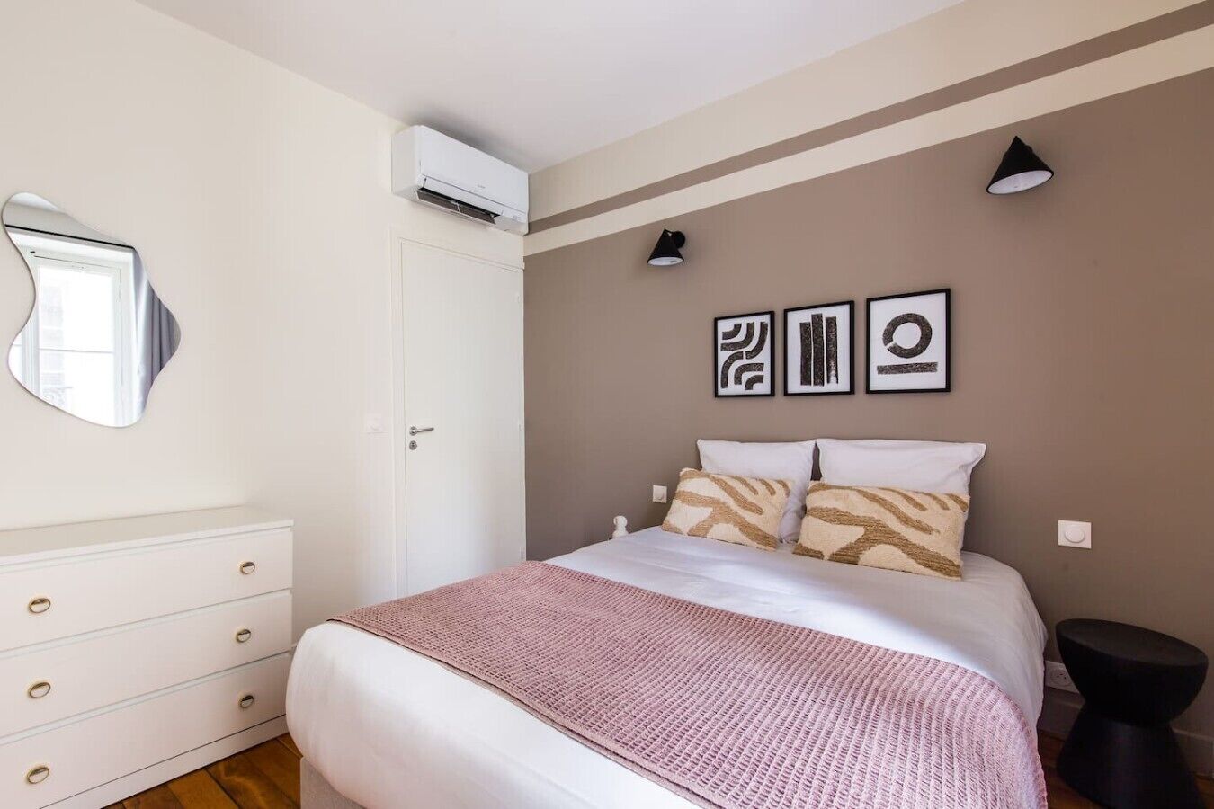 Een slaapkamer met een bed, een dressoir, een airconditioningapparaat, een spiegel en decoratieve wandkunst.