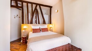 Een slaapkamer met een houten plafond, een bed met witte beddengoed, twee nachtlampjes en een houten bank aan het voeteneind van het bed.