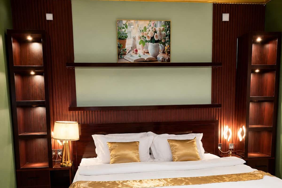 Una cama grande con cubrecama blanco y dorado, flanqueada por cabeceros y estantes de madera, en una habitación con paredes verdes y un techo con iluminación empotrada.