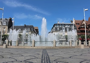 Ein Brunnen mit Wasserstrahlen auf einem öffentlichen Platz, umgeben von Gebäuden im traditionellen Baustil.