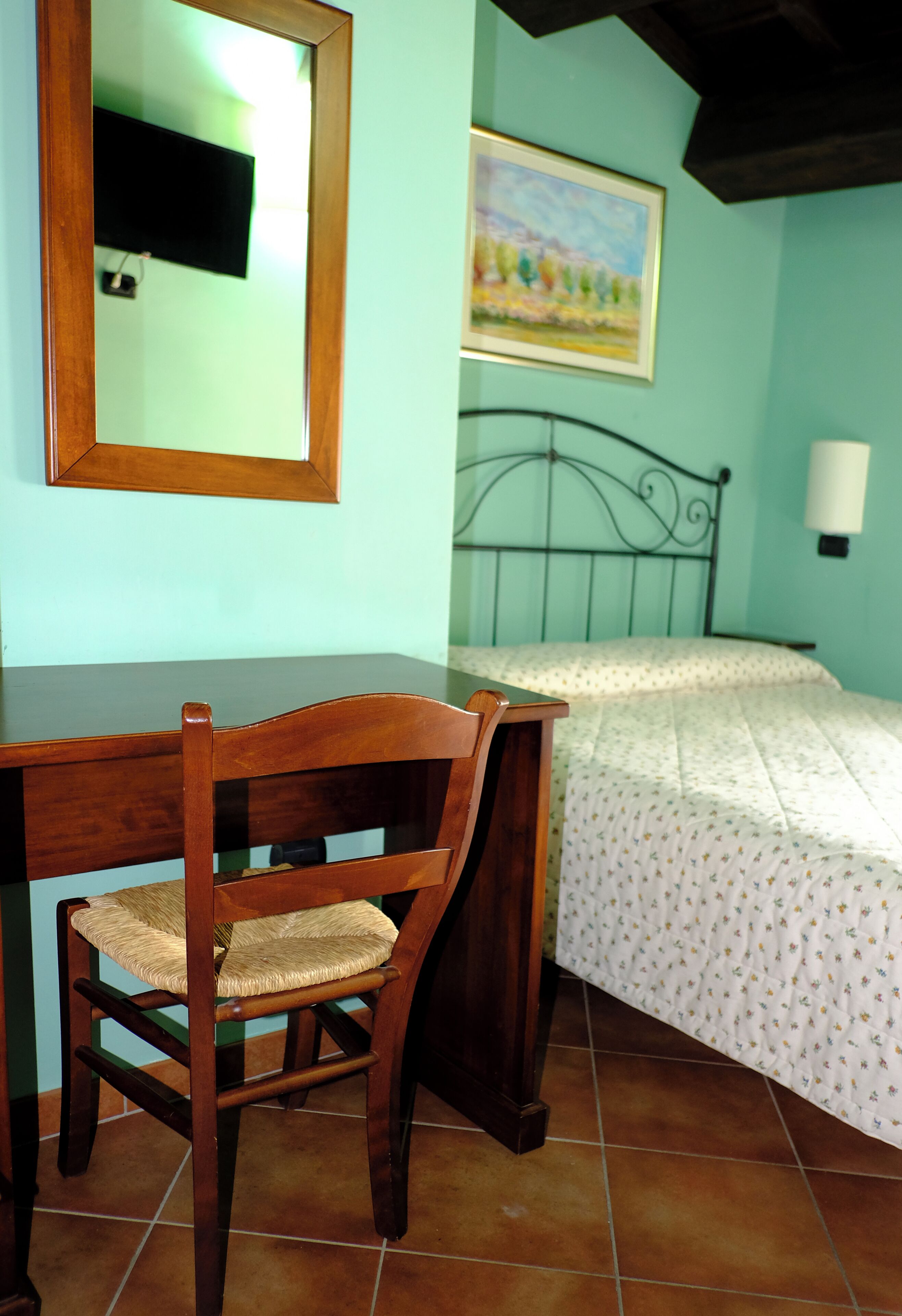 A bedroom with a bed, a wooden chair, a desk, a mirror, and a painting on the wall.