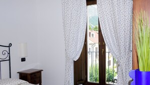 A bedroom with a bed, a nightstand, a wooden chair, a window with curtains, and a view of a balcony.