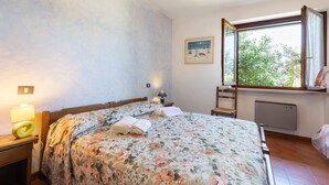 A bedroom with a bed, a nightstand, a lamp, a window with a view, and a radiator.