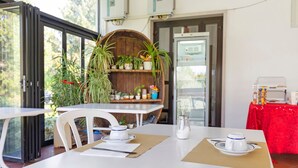 Comedor con una mesa blanca, sillas y mantel rojo. Hay plantas en macetas y una repisa de madera con varios objetos.