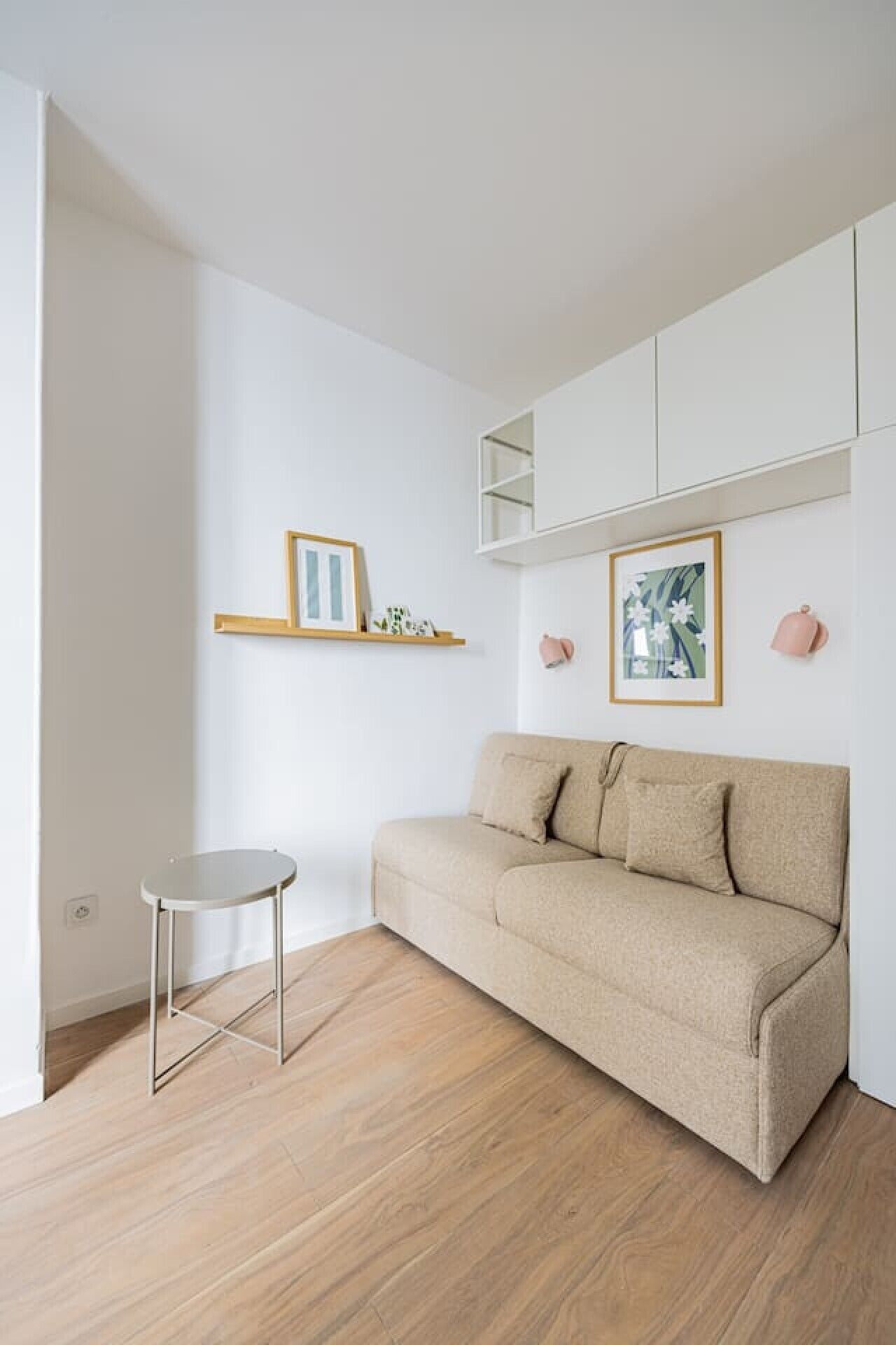 A minimalist living room with a beige sofa, a small round table, and two wall-mounted shelves with framed pictures.