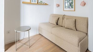 Ett minimalistiskt vardagsrum med en beige soffa, ett litet runt bord och två vägghängda hyllor med inramade bilder.