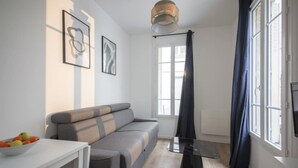 A modern living room with a sofa, a table with fruit, a TV, and a window with curtains. - 5#Studio#2 People#Paris 15#Retail#AC (Paris)
