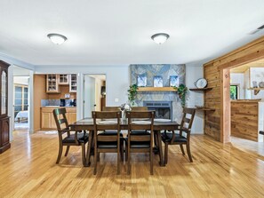 A dining area with a wooden table and chairs, a stone fireplace, and a kitchen in the background. - Sweet Caroline Retreat by Chatuge Home Concierge | Private Pool, hot tub, game room, corn hole, outdoor fireplace, Sauna separate office/yoga studio (Hayesville)