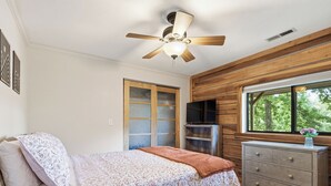 Quarto com cama, penteadeira, ventoinha no teto, televisão e janela com vista para árvores.