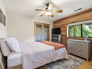 Ein Schlafzimmer mit Bett, Kommode, Deckenventilator, Fernseher und einem Fenster mit Blick auf Bäume.