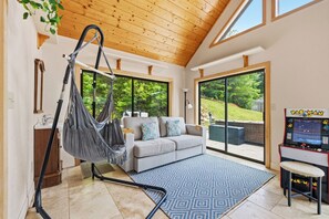 A room with a hammock chair, a sofa, a patterned rug, and a Pac-Man arcade game. - Sweet Caroline Retreat by Chatuge Home Concierge | Private Pool, hot tub, game room, corn hole, outdoor fireplace, Sauna separate office/yoga studio (Hayesville)