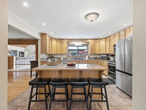 A modern kitchen with wooden cabinets, a central island with stools, stainless steel appliances, and a window. - Sweet Caroline Retreat by Chatuge Home Concierge | Private Pool, hot tub, game room, corn hole, outdoor fireplace, Sauna separate office/yoga studio (Hayesville)