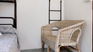 Una habitació amb un llit, una cadira de vimet i una escala metàl·lica adossada a la paret.