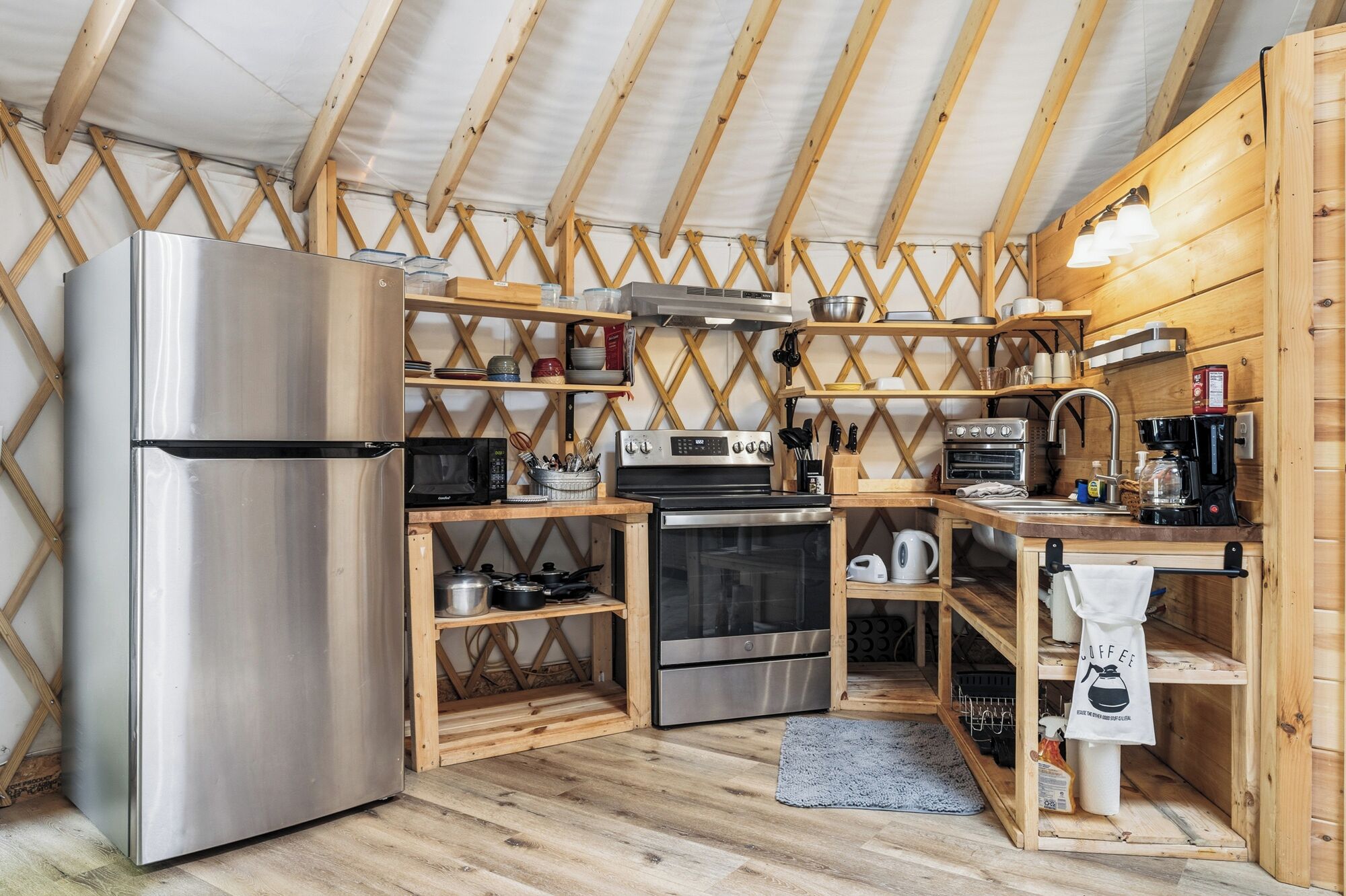 A compact kitchen with a stainless steel refrigerator, microwave, stove, oven, and coffee maker. Wooden shelves and a wooden ceiling are visible.
