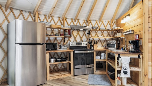 Eine kompakte Küche mit Edelstahl-Kühlschrank, Mikrowelle, Herd, Backofen und Kaffeemaschine. Holzregale und eine Holzbalkendecke sind zu sehen.