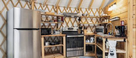 A compact kitchen with a stainless steel refrigerator, microwave, stove, oven, and coffee maker. Wooden shelves and a wooden ceiling are visible.