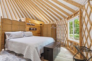 Ein gemütlicher, hell erleuchteter Zeltraum mit Bett, Bank, einer Kleiderstange und einem Fenster mit Blick ins Grüne.