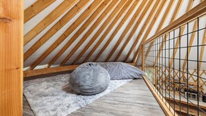 Un dormitorio tipo loft con techo inclinado, vigas de madera, un otoman tejido color gris y una alfombra blanca y mullida.