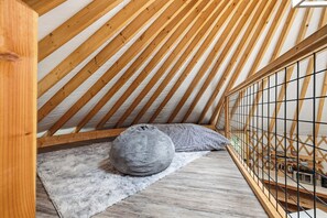 A loft bedroom with a sloped ceiling, wooden beams, a grey knitted pouf, and a white furry rug. - The Wanderer's Yurt #1 by Chatuge Home Concierge | Luxury Mountain Glamping (Hayesville)