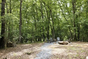 A forested campsite with a gravel path, wooden benches, and a pile of logs. - The Wanderer's Yurt #1 by Chatuge Home Concierge | Luxury Mountain Glamping (Hayesville)