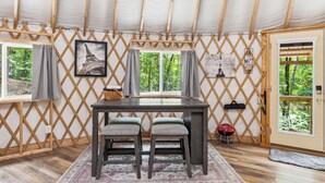 Habitación con carpa, una mesa, dos sillas, una ventana con cortinas, una puerta y vista a árboles.